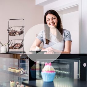 A person stands behind a pastry counter in a bakery setting, with various decorated cupcakes and desserts displayed in a glass case and additional baked goods arranged in wire baskets on the counter.